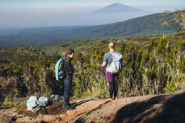 Woman and man with backpacks relaxing after the hiking trip on Mount Kilimanjaro looking at the beautiful blue mountain and the green valley